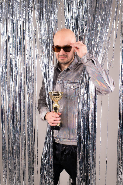 Strange caucasian man in a glittering white shirt, black leather pants, grey shoes and black sunglasses received a gold cup. - Фото, изображение