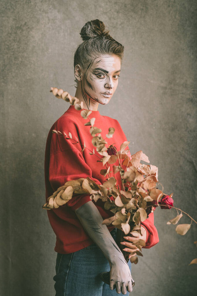 Beautiful young woman with creative drawings one her face (cartoon skeleton) holding vase with dry decorative flowers over grunge wall on background. - Фото, зображення
