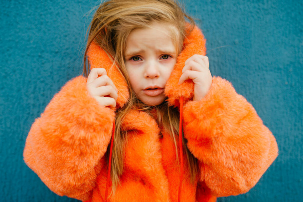 A beautiful little girl with medium fair hair in a bright orange fur coat stands near the long blue wall. - Foto, Bild