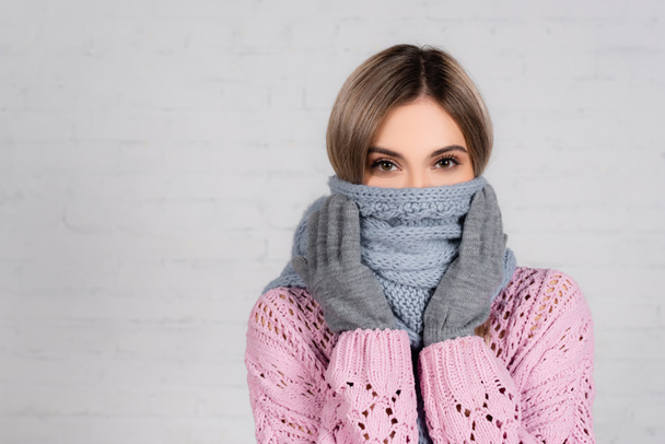 Woman in knitted sweater and gloves wrapped in scarf looking at camera on white background - 写真・画像