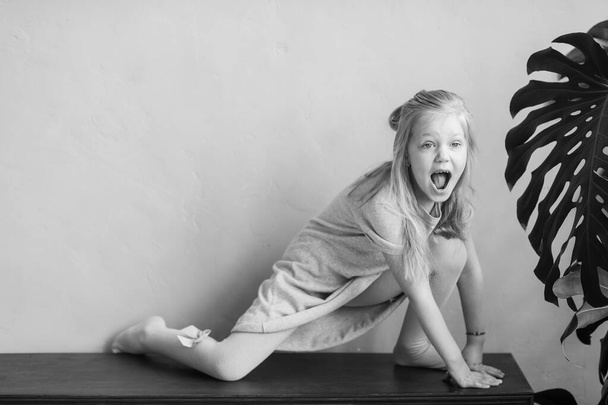 Beautiful little blonde long haired child girl in white tights posing for camera with excited funny face behind wall. Comical female child make faces and having fun in studio. Happy kid bw portrait - Zdjęcie, obraz