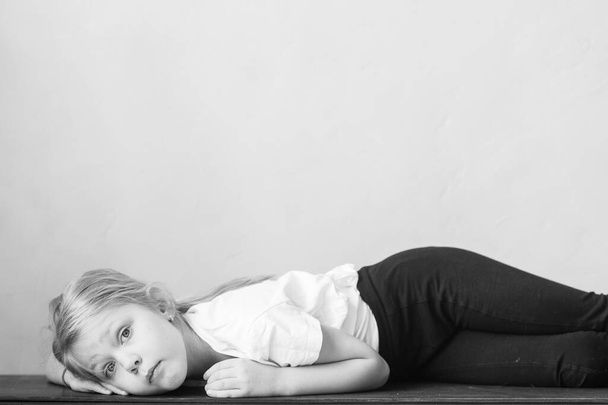 Lonely little girl with pensive tranquil face lying on wooden surface at home and looking at camera with deep emotional eyes.  Lifestyle portrait of poor offensed female child on wall background - Photo, Image
