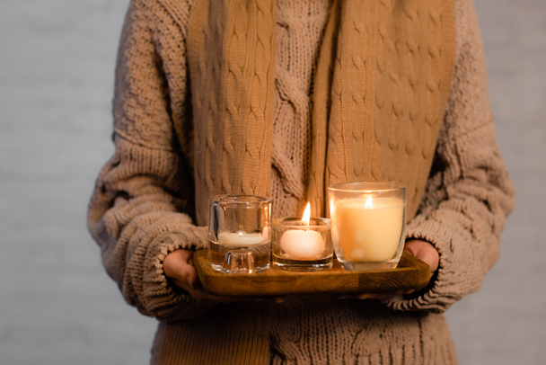 Cropped view of burning candles on wooden board in hands of woman in scarf and sweater blurred on white background - Photo, Image