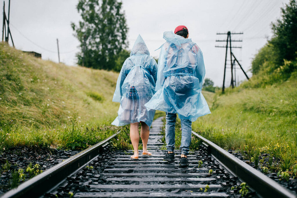 Two young travelers in rain cloaks go on outgoing railway overgrown with grass. Company of friends rest on vacation. Tourist clothes. Cloudy muggy weather. Hipster lifestyle. Rails in perspective - Photo, Image