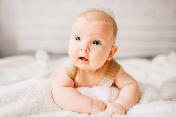 Adorable lovely newborn male baby boy with smiling emotional  happy face lifestyle indoor portrait. Funny infant child lying on stomach on bed with white wall on background.  Carefree childhood - Zdjęcie, obraz