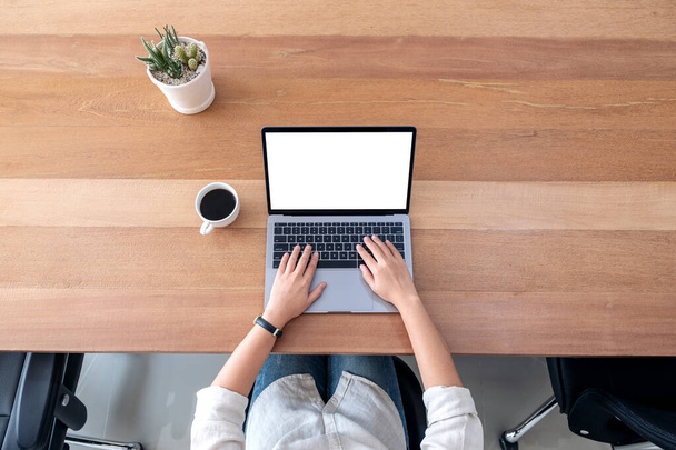 オフィスの木製テーブルの上に空白の白いデスクトップ画面とラップトップを使用して入力する女性のトップ表示モックアップイメージ - 写真・画像