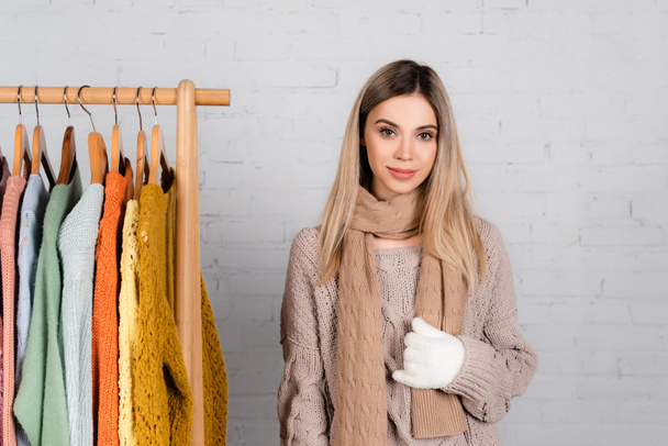 Lächelnde Frau in Strickwaren und Handschuhen, die in der Nähe von Kleiderständern mit Pullovern auf weißem Hintergrund steht - Foto, Bild