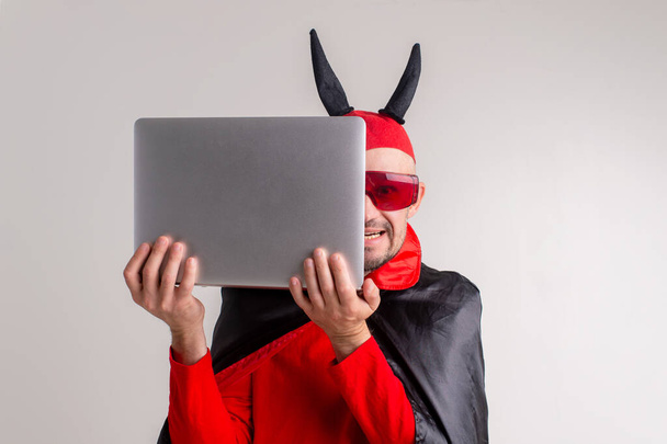 Stylish caucasian man in devil hat with horns and vampire cape with laptop isolated on white backhround. - Fotó, kép