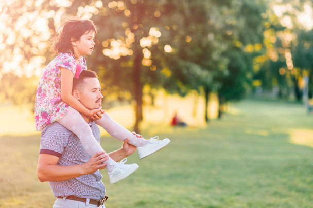 Dad carrying his daughter on shoulders in summer park at sunset. Father have fun with his little child. Kid siiting at parent neck. Happy family playing and making  crazy faces.  Lifestely portrait. - 写真・画像