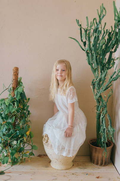 Beautiful  little blonde long haired child girl in white dress standing in clay pot between two green plants on decorated yellow wall background. Funny cheerful female kid with surprised face portrait - Photo, image