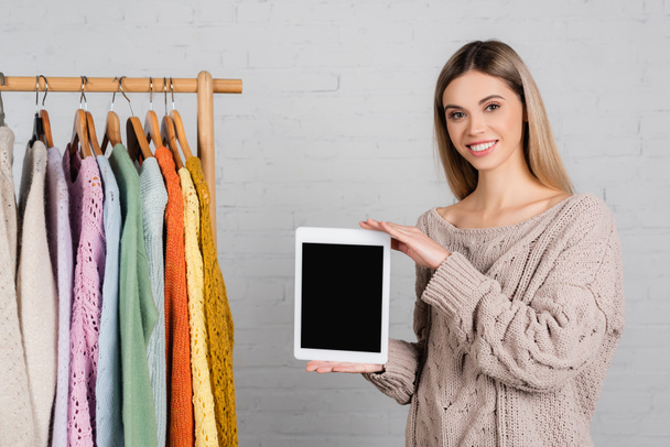 Mosolygó nő gazdaság digitális tabletta üres képernyőn közelében fogas állvány pulóverek fehér háttér - Fotó, kép
