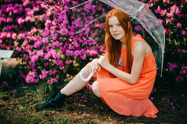 Strange young girl with long red hair, pretty face in a summer orange dress holds an umbrella and have a good time in the garden. - Фото, изображение