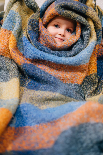 Pretty caucasian little girl with fair hair in blue dress sits in baby room wrapped herself in a colourful coverlet and smiles - 写真・画像