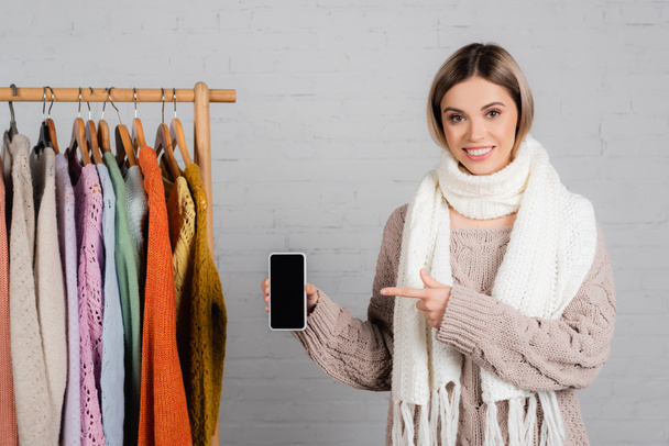 Smiling woman pointing at smartphone with blank screen near hanger rack with sweaters on white background - 写真・画像