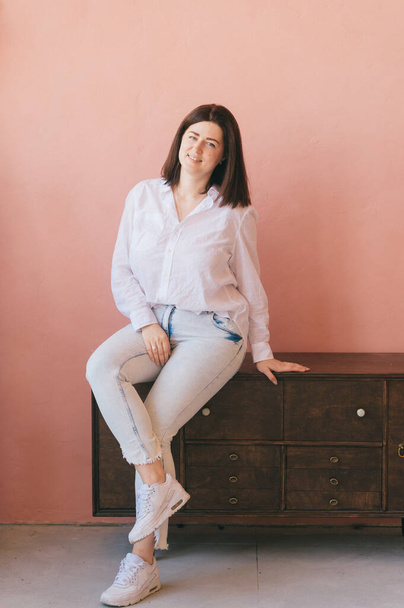Beautiful brunette young woman relaxing and posing for camera on pink wall backgrorund. Lifestyle indoor portrait of cheerful smiling happy adult caucasian female sitting on wooden commode at home - Φωτογραφία, εικόνα