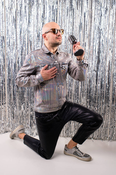 A man without hair with sunglasses in a shiny shirt, black leather pants and grey sneakers sings in a small silver microphone. - Fotoğraf, Görsel