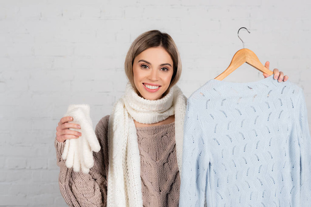 Smiling woman in scarf holding hanger with sweater and gloves on white background - Zdjęcie, obraz