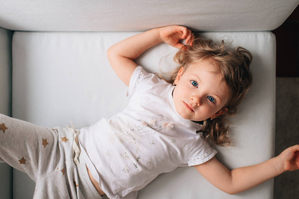 Portrait of cute little caucasian girl in white sleepwear lies on her white bed and rejoices - Foto, imagen