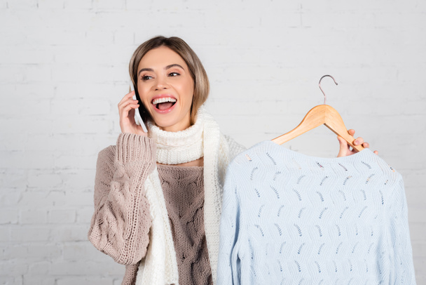 Mujer alegre en ropa de abrigo hablando en el teléfono celular y sosteniendo percha con suéter sobre fondo blanco - Foto, Imagen