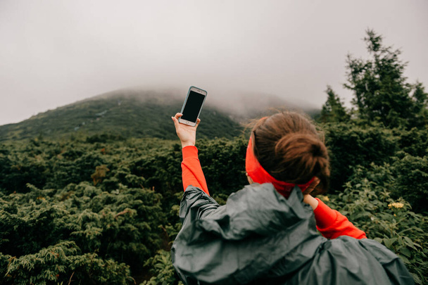 Goverla, Ukraine - July 14, 2018: Lifestyle portrait of adventure traveler girl in wet raincoat take selfie on smartphone high in foggy mountains under rain. Young female hiker take self pic on phone  - Foto, afbeelding