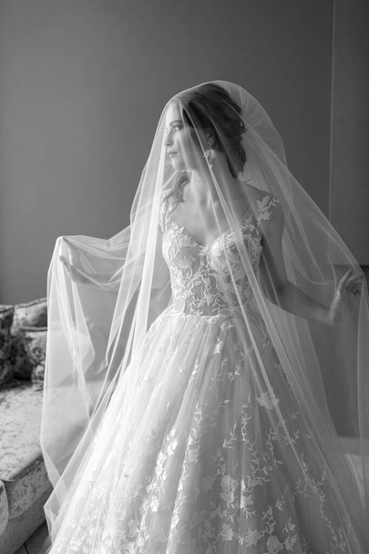 Black and white portrait of beautiful bride in wedding dress touchs and throws her veil up in hotel. - Fotó, kép