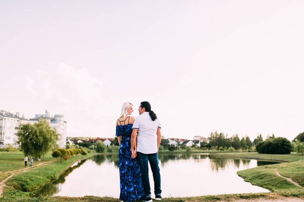 long-haired man and slender tall blonde in long blue dress look at magnificent lake on a Sunny summer day. Love story of guy and girl overlooking the river. Lovers on background of beautiful landscape - Photo, Image