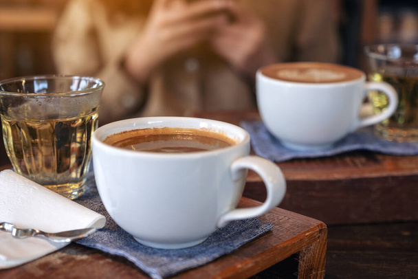Closeup image of two white cups of hot coffee and a glass of tea on vintage wooden table with a woman in cafe - Zdjęcie, obraz