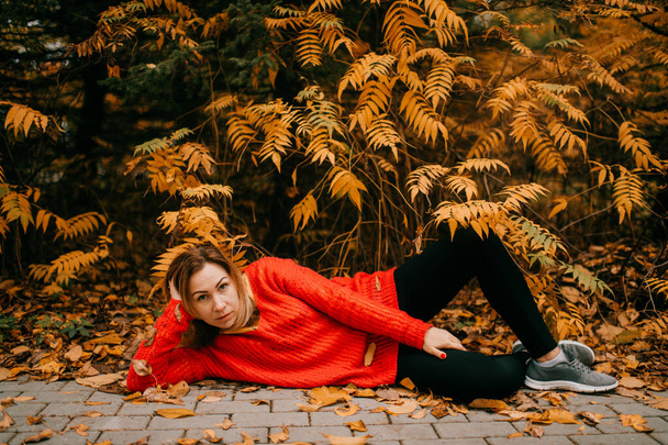Portrait of charming caucasian female in a red sweater, black pants and colourful knitting hat lies on the yellow grass and relaxes after hard day. - Foto, Imagen