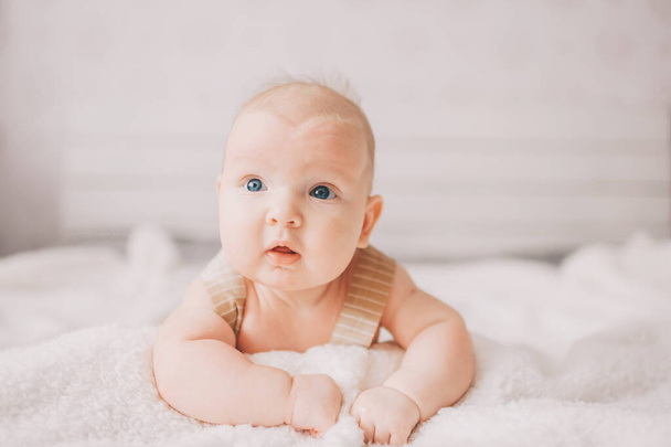 Adorable lovely newborn male baby boy with smiling emotional  happy face lifestyle indoor portrait. Funny infant child lying on stomach on bed with white wall on background.  Carefree childhood - Foto, Imagen