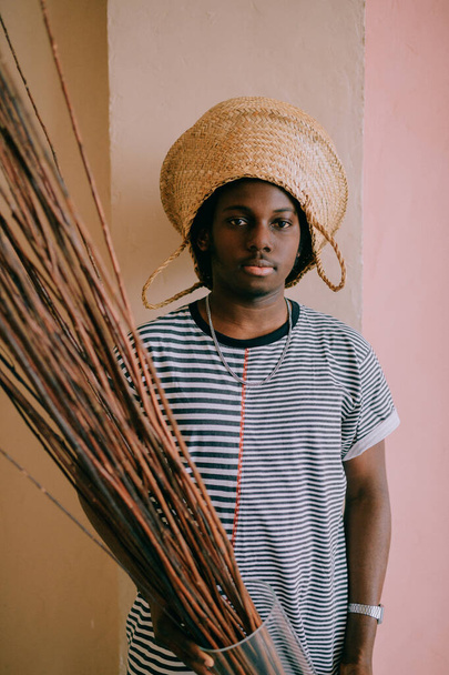 Lifestyle portrait of strange young african black man holding glass vase with wooden twigs in hands and straw basket on his head. Dark-skinned nigerian male indoor funny  portrait on pink wall at home - Photo, Image