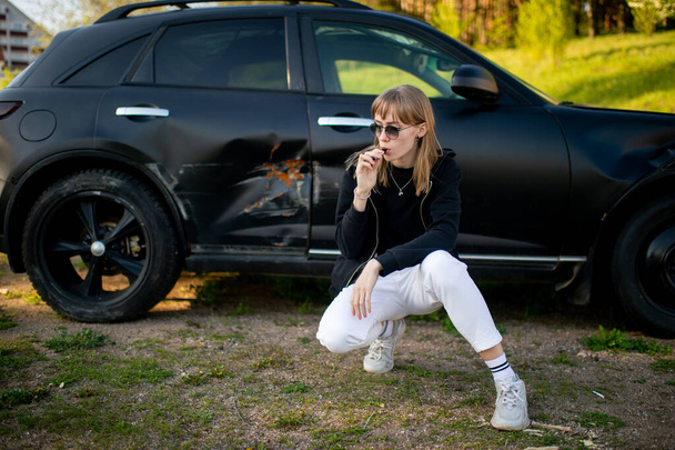 Beautiful young caucasian woman in white jeans, white trainers and black jacket poses for the camera with old black briken car outside. - Foto, immagini