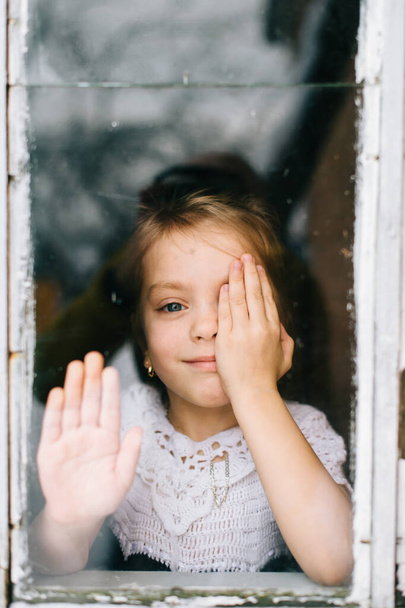 Vidám mosolygós lány eltakarja a szemét egy kézzel, és a kamerát nézi az ablakon keresztül elvont gondolatok. - Fotó, kép