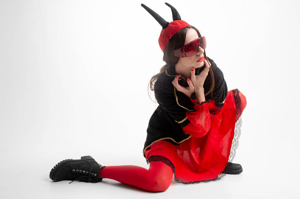 Sexy brunette girl in red tights, masquerade costume, futuristic glasses and devil horns on her head posing with passion over white background - Foto, imagen