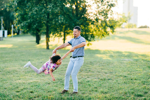 Father holding his daaughter hand with leg and whirling around. Excited family have fun in summer park. Dad playing with little female child on green meadow. Carefree happy childhood. Fatherhood  - Foto, Imagem