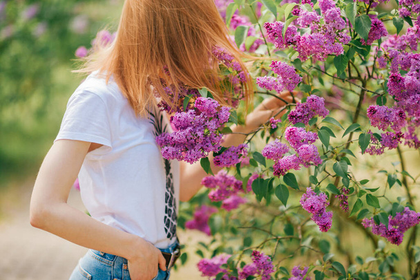 Beautiful young caucasian woman with red hair turned away in lilac garden with flowers - Φωτογραφία, εικόνα