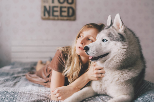 Young girl in brown dress lying on bed at home and sleeping with husky puppy. Lifestyle indoor portrait of beautiful woman hugs husky dog on sofa. Pet lover. Cheerful female resting with adorable dog - Фото, зображення