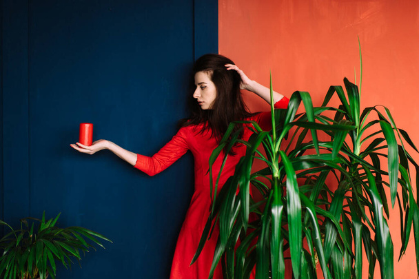 Beautiful fashion girl with dark long hair, spanish appearance, in red elegant dress holding candle on blue red wall. Indoor soft focus portrait of brunette woman showing trick like magician - Photo, image