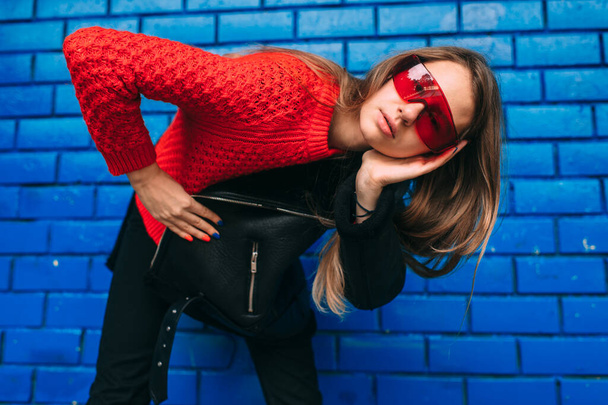 Fashion portrait of young long haired brunnete woman in black sheepskin coat, red sportive sunglasses posing over blue brick wall outdoors. - Fotografie, Obrázek
