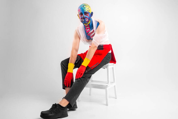 Dziwny mężczyzna z makijażem na głowie i ramieniu w białej koszulce, czerwonych i żółtych rękawiczkach, czerwony siedzi na białym krześle - Zdjęcie, obraz