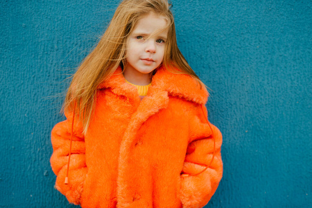 Ένα όμορφο κοριτσάκι με μεσαία ξανθά μαλλιά σε ένα φωτεινό πορτοκαλί γούνα στέκεται κοντά στο μακρύ μπλε τοίχο. - Φωτογραφία, εικόνα