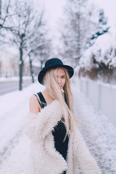Stijlvol mooi blond meisje in jas en hoed poseren in de buurt hek in de straat - Foto, afbeelding