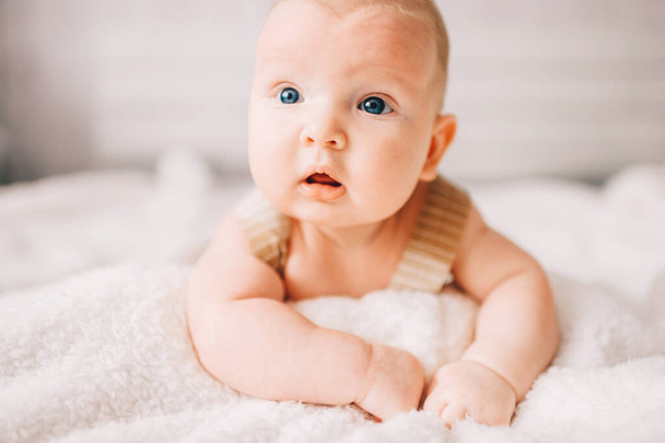 Adorable lovely newborn male baby boy with smiling emotional  happy face lifestyle indoor portrait. Funny infant child lying on stomach on bed with white wall on background.  Carefree childhood - Foto, Imagem