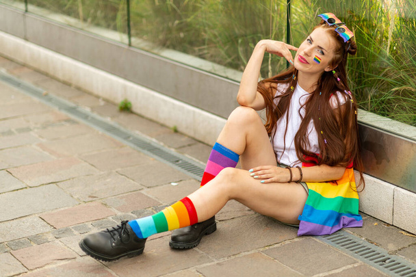 Stylish lesbian model with lgbt flag on her face sitting near glass wall outdoor  - Φωτογραφία, εικόνα