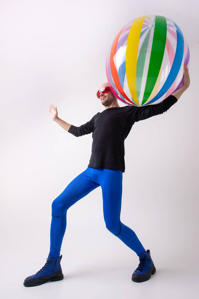 Fitness young man in blue sportive tights, black athletic shirt and stylish boots holding beach ball on his houlder against white studio background - Foto, Imagem