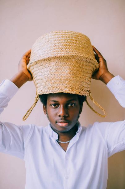 Closeup lifestyle portrait of strange young african black man holding straw basket above his head on textured wall background and looking at camera. Dark-skinned boy in white t-shirt funny portrait. - Photo, Image