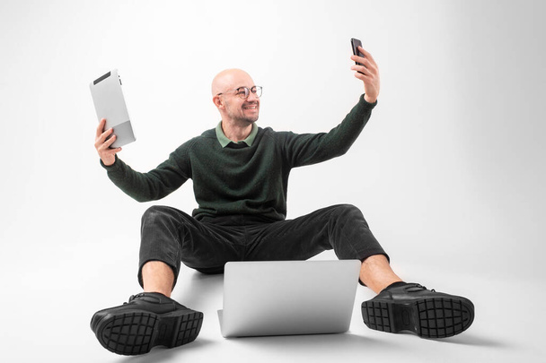 Bonito homem caucasiano segura um telefone em uma mão, tablet em outra mão com laptop, senta-se em um piso do selfie isolado no fundo branco - Foto, Imagem