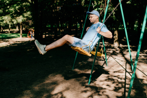 Adult stylish man in glasses riding on swing in city park on playground for children in summer.  Happy guy remember childhood.  Male person on attraction enjoying up and down motion.  Odd boy outdoor - Foto, imagen