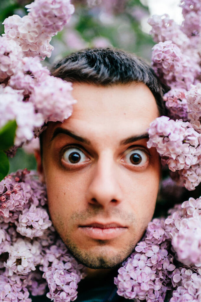 Closeup odd unusual male portrait outdoor at nature in beautiful lilac bushes.  Adult funny man face grimace surrounded by blooming flowers. Emotions and facial expxression. Clowning and fooling - Photo, Image