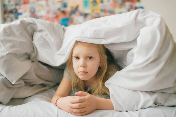Young adorable blonde girl with funny face lying on bed under white blanket and look at camera with sad eyes. Little female child with pensive face indoor lifestyle portrait. Little  girl portrait - Photo, Image