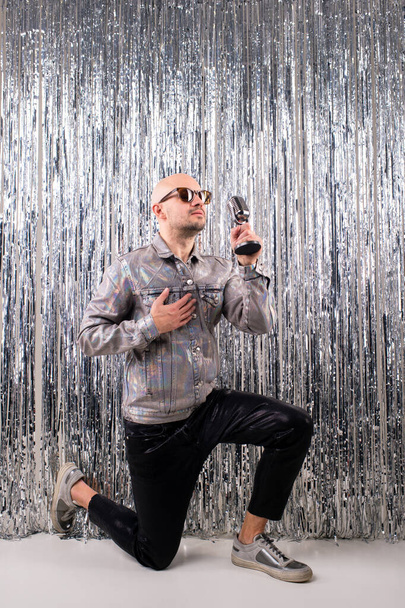 Picture of beautiful caucasian man without hair with black sunglasses in a shiny shirt, black leather pants and grey sneakers sings in a small silver microphone isolated on silver glitter background. - Photo, Image
